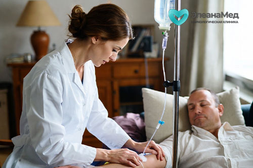 Врач-нарколог в белом халате ставит капельницу мужчине 40 лет на диване в домашней обстановке